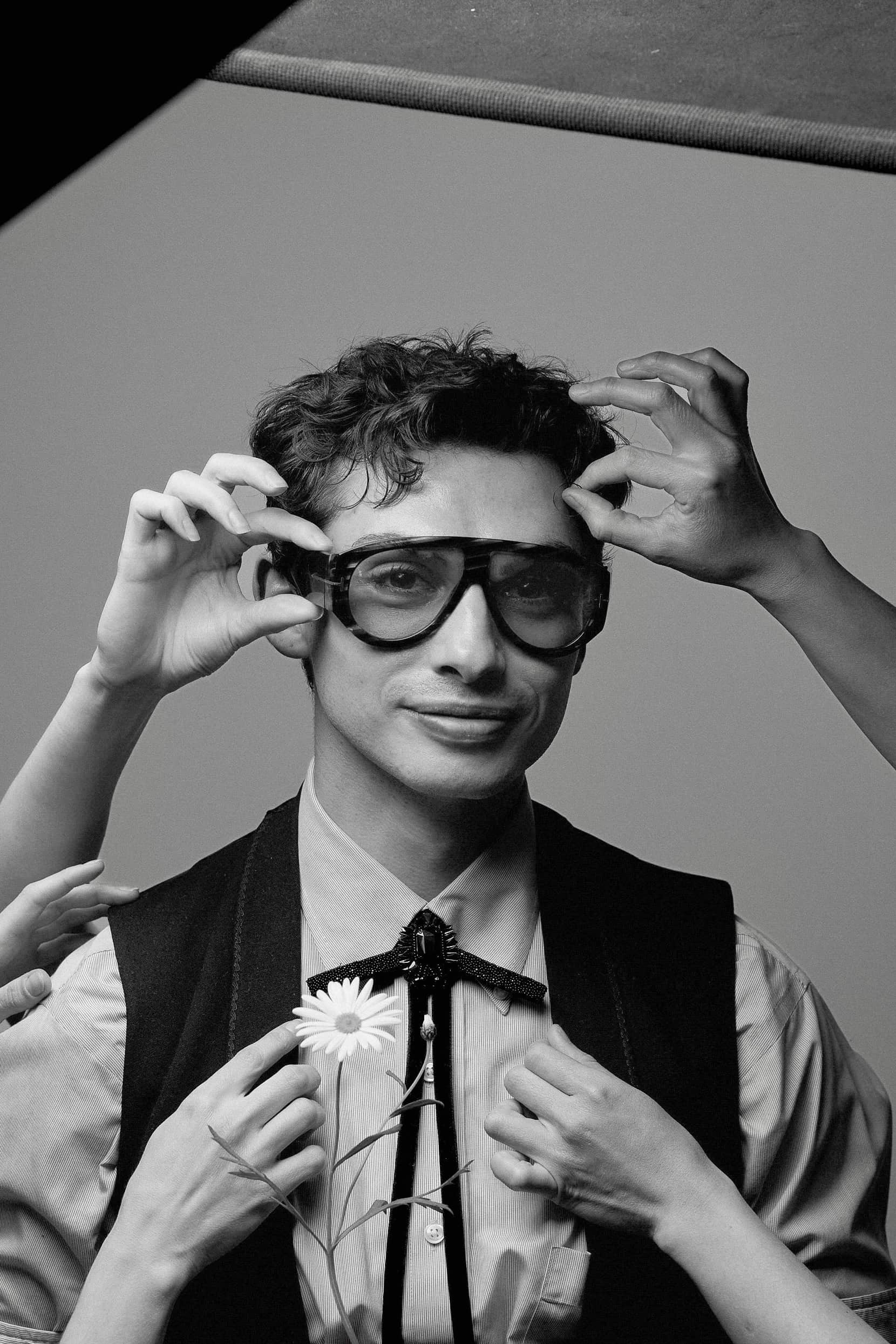 A portrait of Nicholas Alvoeiro, founder of Act II. He has dark hair and is wearing a grey shirt, black vest, a bolo tie, and sunglasses. Hands reach from out of frame to adjust his sunglasses and vest.